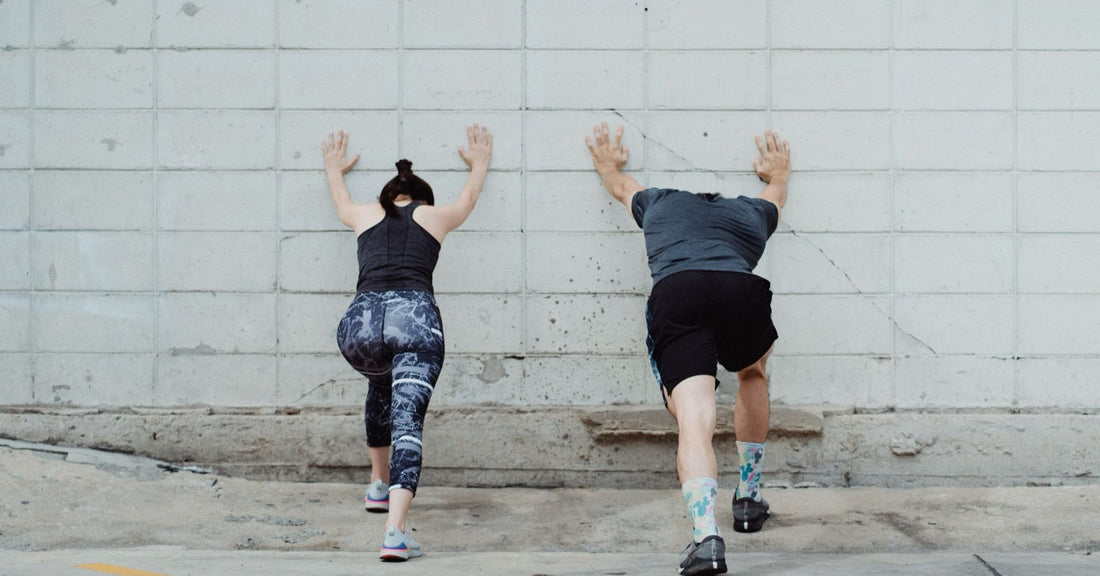 Two people leaning against a concrete wall doing a standing calf stretch, back heels pressed down.
