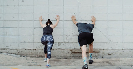 Two people leaning against a concrete wall doing a standing calf stretch, back heels pressed down.