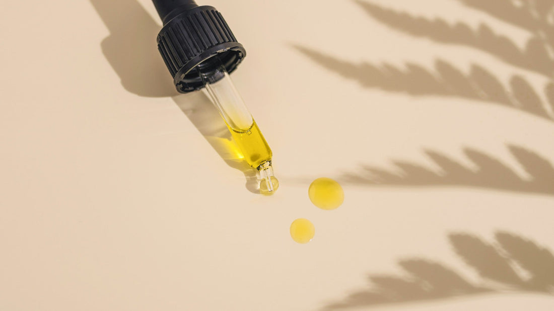 Glass dropper releasing drops of golden hemp oil beside the shadow of a hemp leaf.